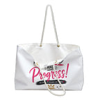 A white canvas tote bag with white rope handles, featuring a graphic print on the front that reads 'AND Progress!' in pink and black, with a small gold crown logo at the bottom.