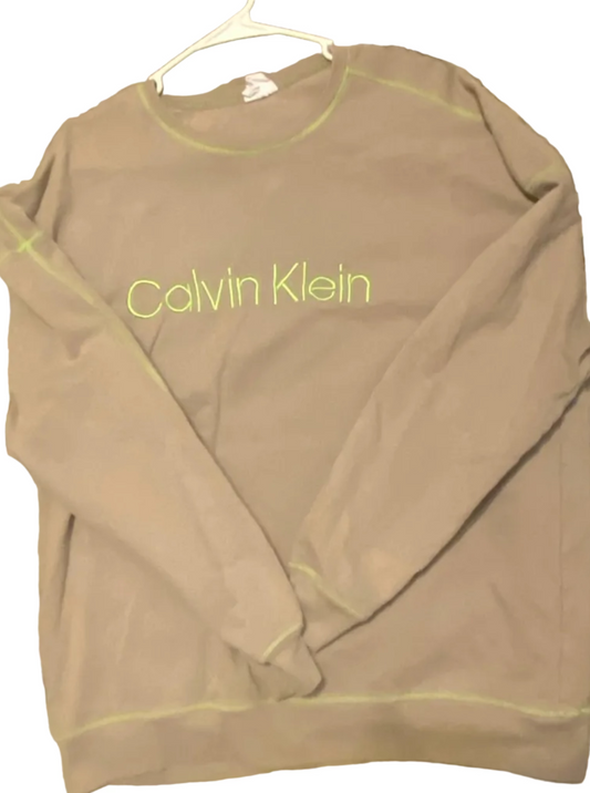 A Calvin Klein long-sleeve athletic top in taupe brown, laid flat on a concrete floor with a white hanger. The shirt features the brand name printed vertically in lime green on the chest and has matching lime green piping along the edges.