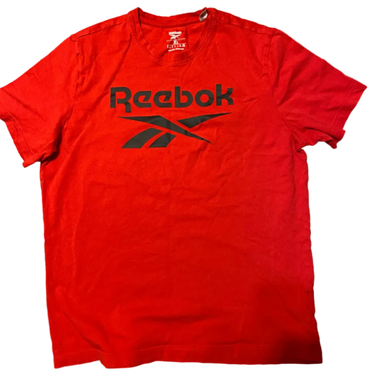A red Reebok short-sleeve t-shirt with a black logo print, laid flat on a textured concrete floor.