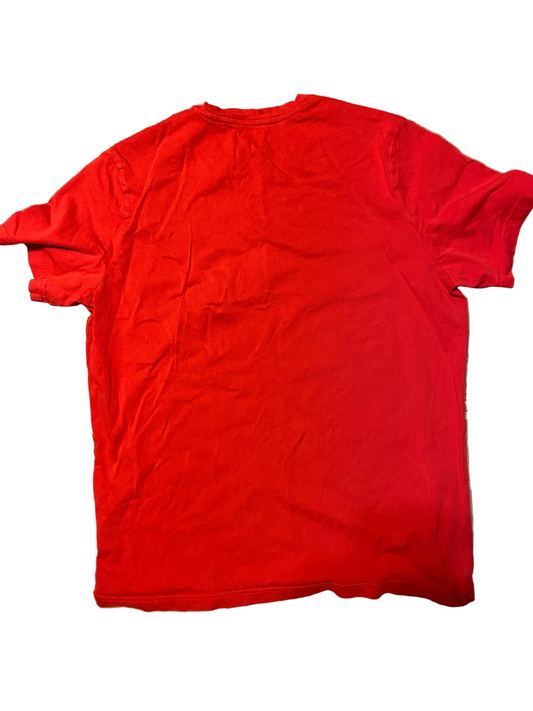 A bright red short-sleeve crewneck t-shirt laid flat on a textured gray and beige patterned floor, with parts of black sandals visible at the bottom corners.