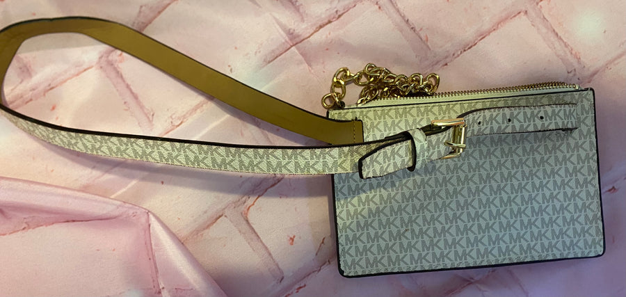 A Michael Kors crossbody wallet in light gray monogram canvas with a gold chain strap and a tan leather wristlet strap, displayed on a pink marble-patterned background.