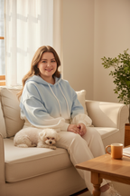 Curvy woman wearing gradient blue-to-white oversized hoodie sitting on cream couch with fluffy white dog