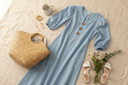 Light blue puff sleeve maxi dress flat lay styled with straw bag, gold jewelry, sandals, and wildflowers on cream linen